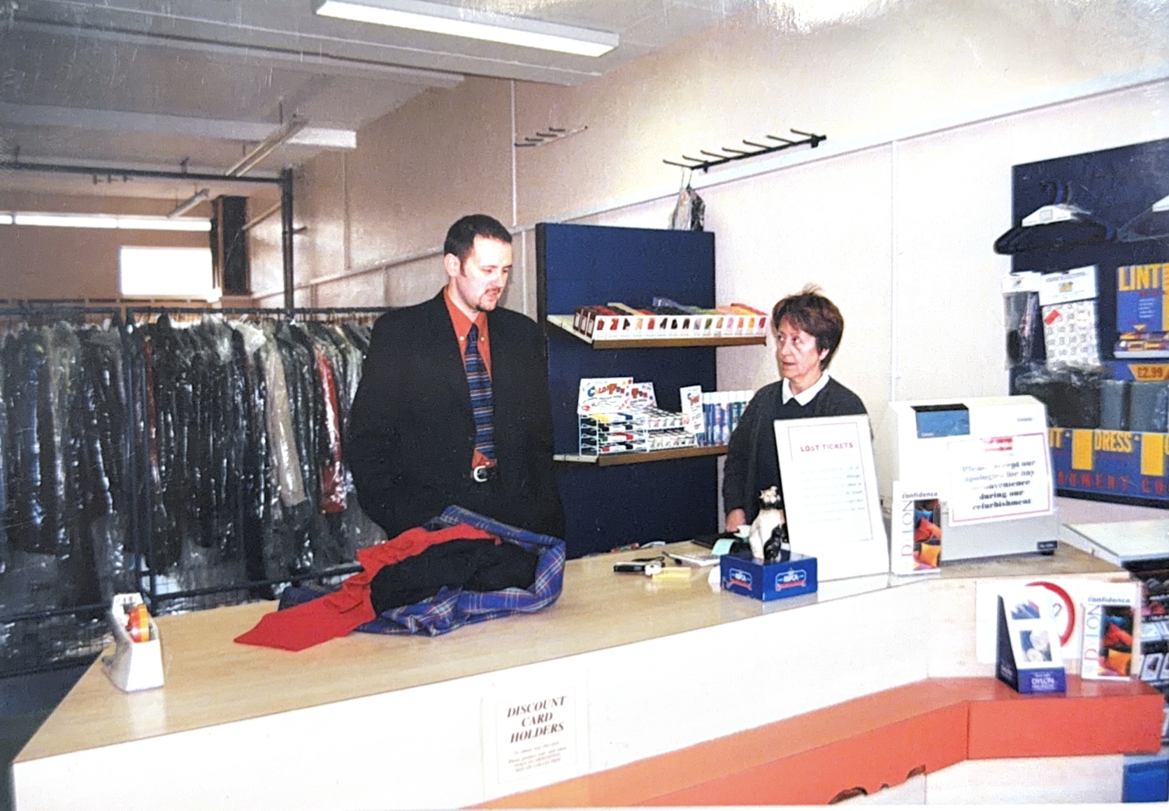 A man and woman stood behind the counter with dry cleaning behind them